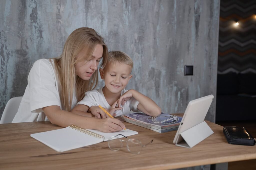 A mother helps her son with homework using a tablet and notebook at a wooden table indoors.